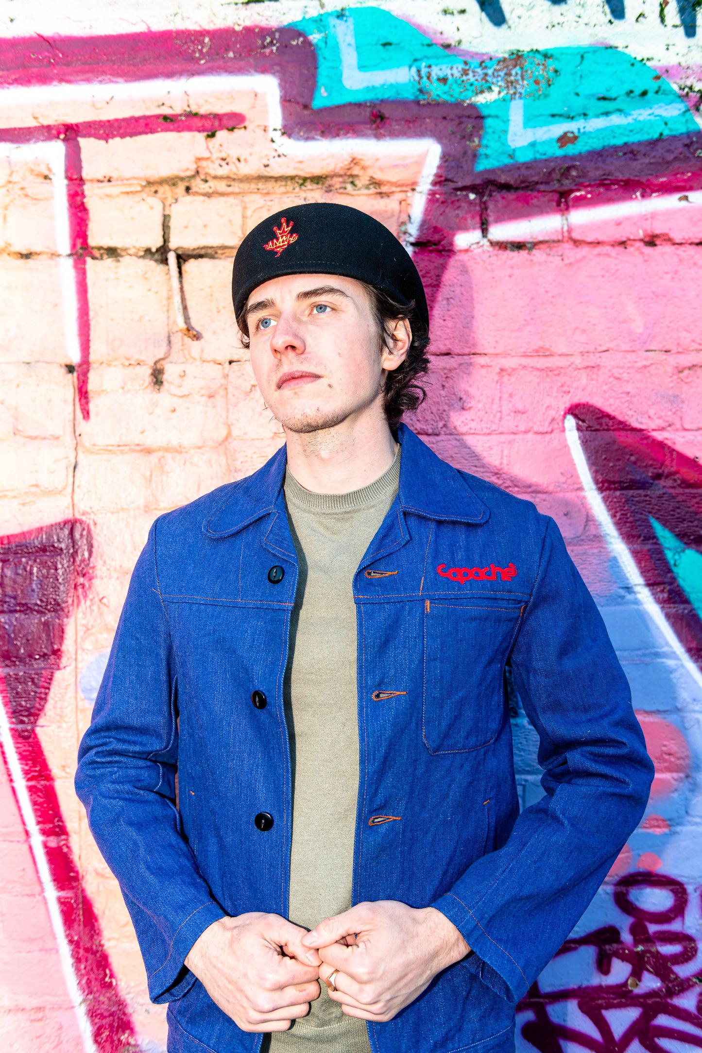 Young man wearing Apache Prison Officer Jacket in rich blue denim with red Capache embroidery, olive crew neck underneath, against colorful graffiti wall