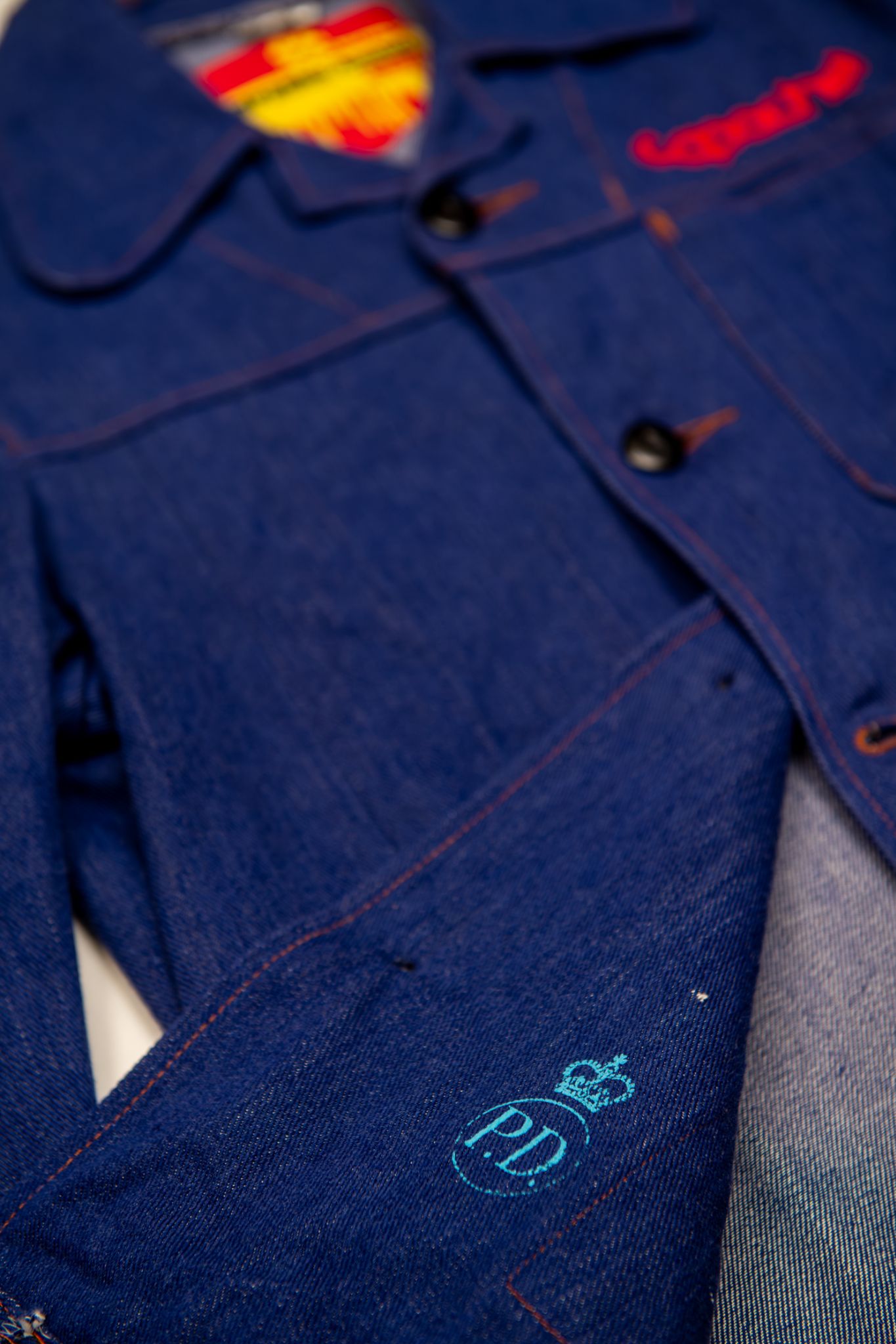 Dark blue denim Apache Prison Officer Jacket with orange contrast stitching, red embroidered details, and teal PD crown logo