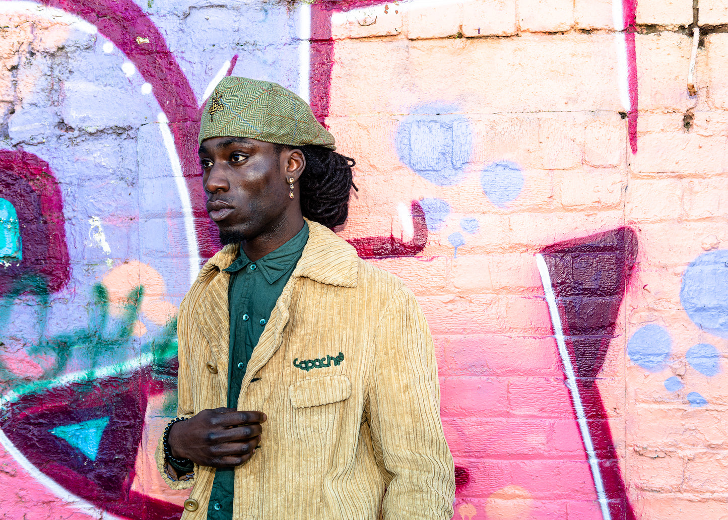 Man wearing Apache Cord Blazer in tan corduroy with green embroidered logo, styled with green button-up shirt and tweed flat cap