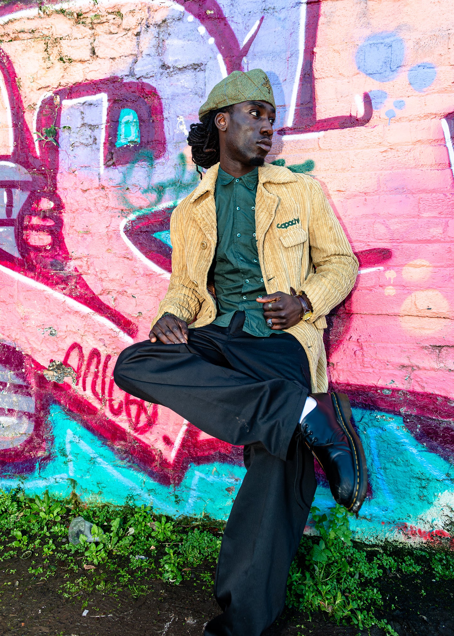 Man wearing Apache Cord Blazer in tan corduroy with green embroidered logo, styled with green shirt, black trousers and flat cap