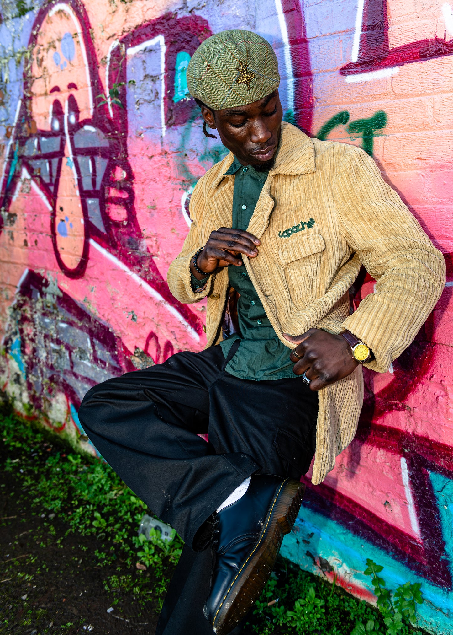 Man wearing tan Apache Cord Blazer with green embroidered logo, styled with green shirt, black trousers and tweed flat cap