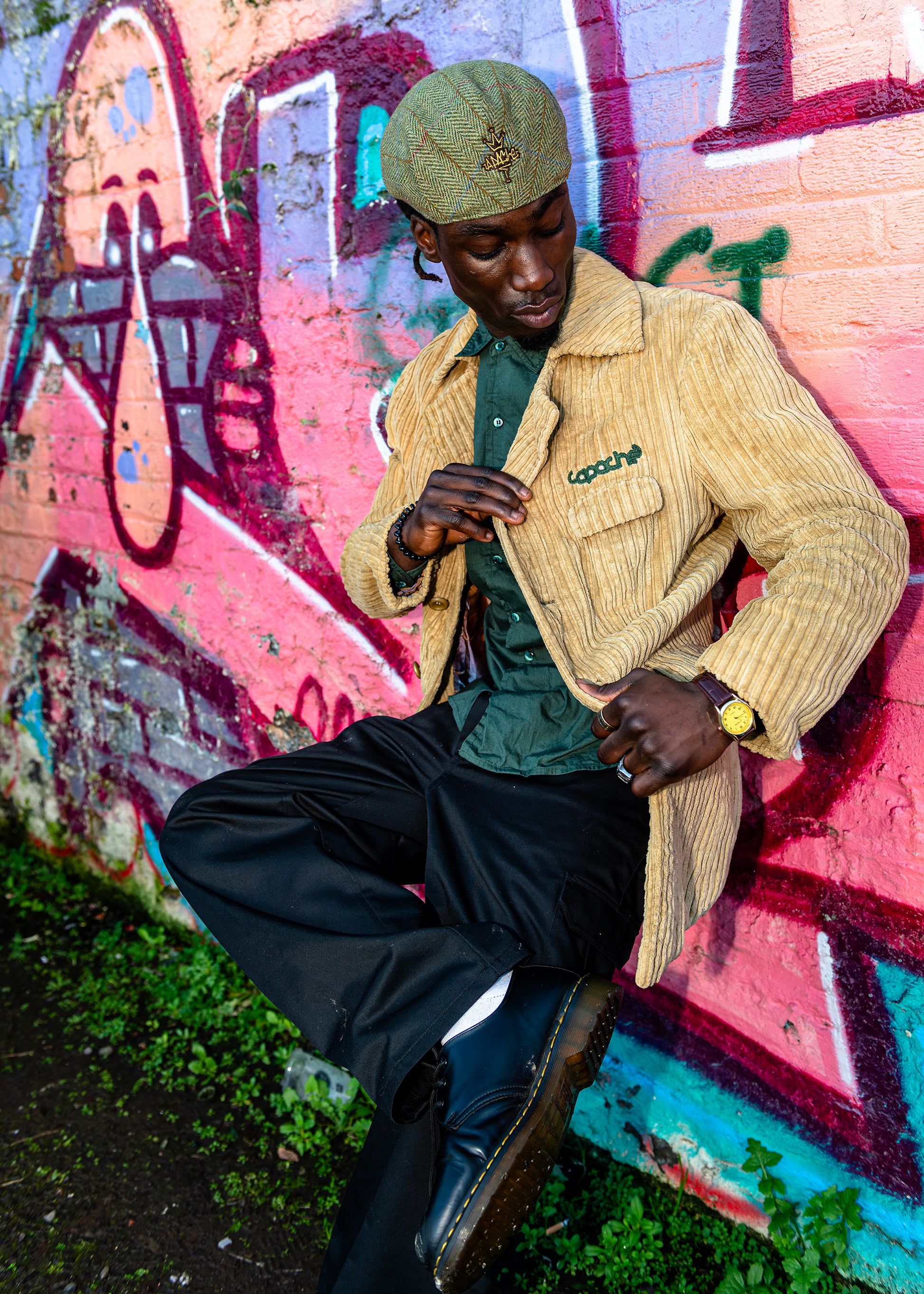 Man wearing tan Apache Cord Blazer with green embroidered logo, styled with green shirt, black trousers and tweed flat cap