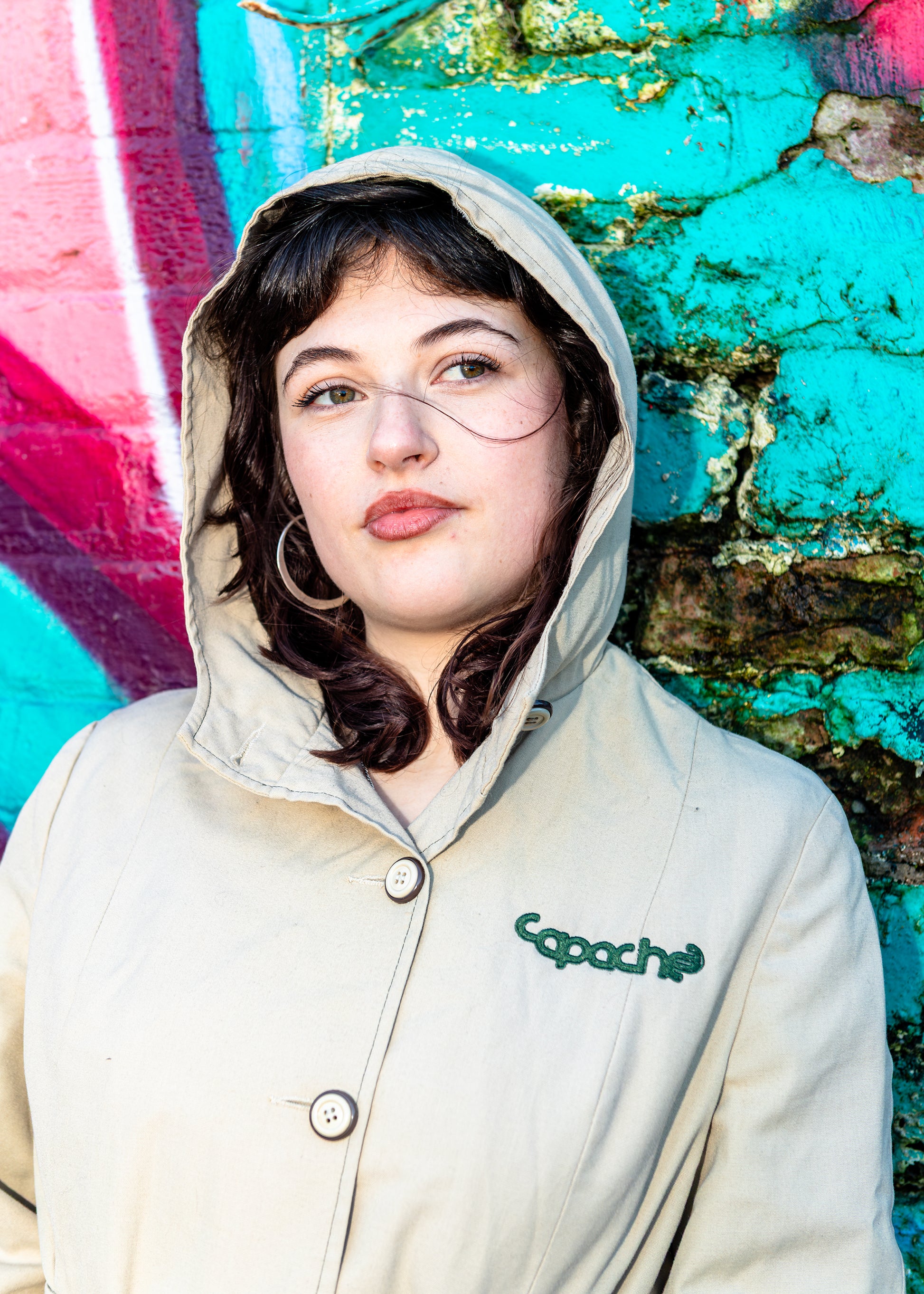Apache Women's Parka in beige with hood, button front closure, and embroidered Capoche logo, styled against colorful graffiti wall