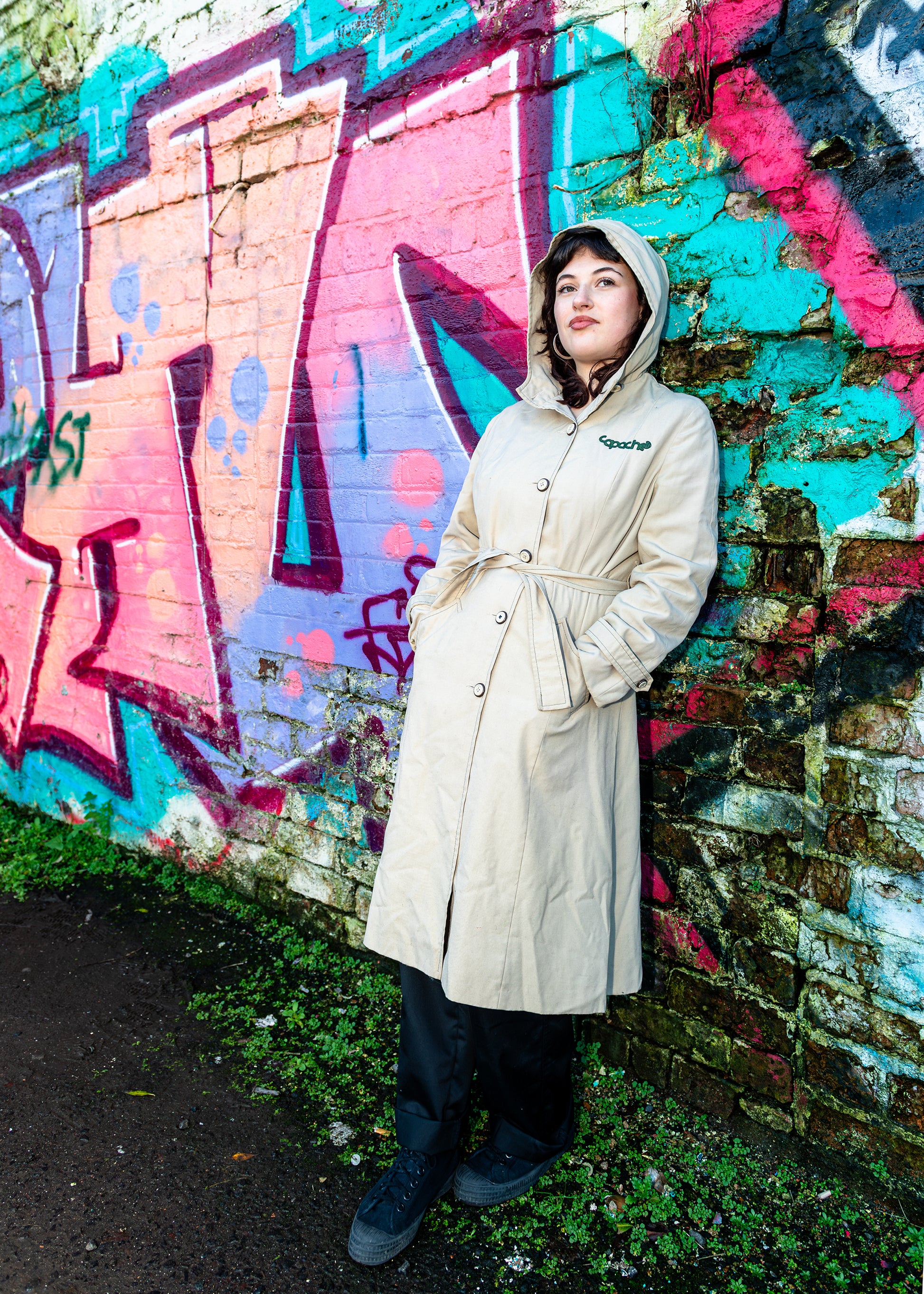 Apache Women's Parka in cream beige with hood, button front and tie waist, worn against colorful graffiti wall background