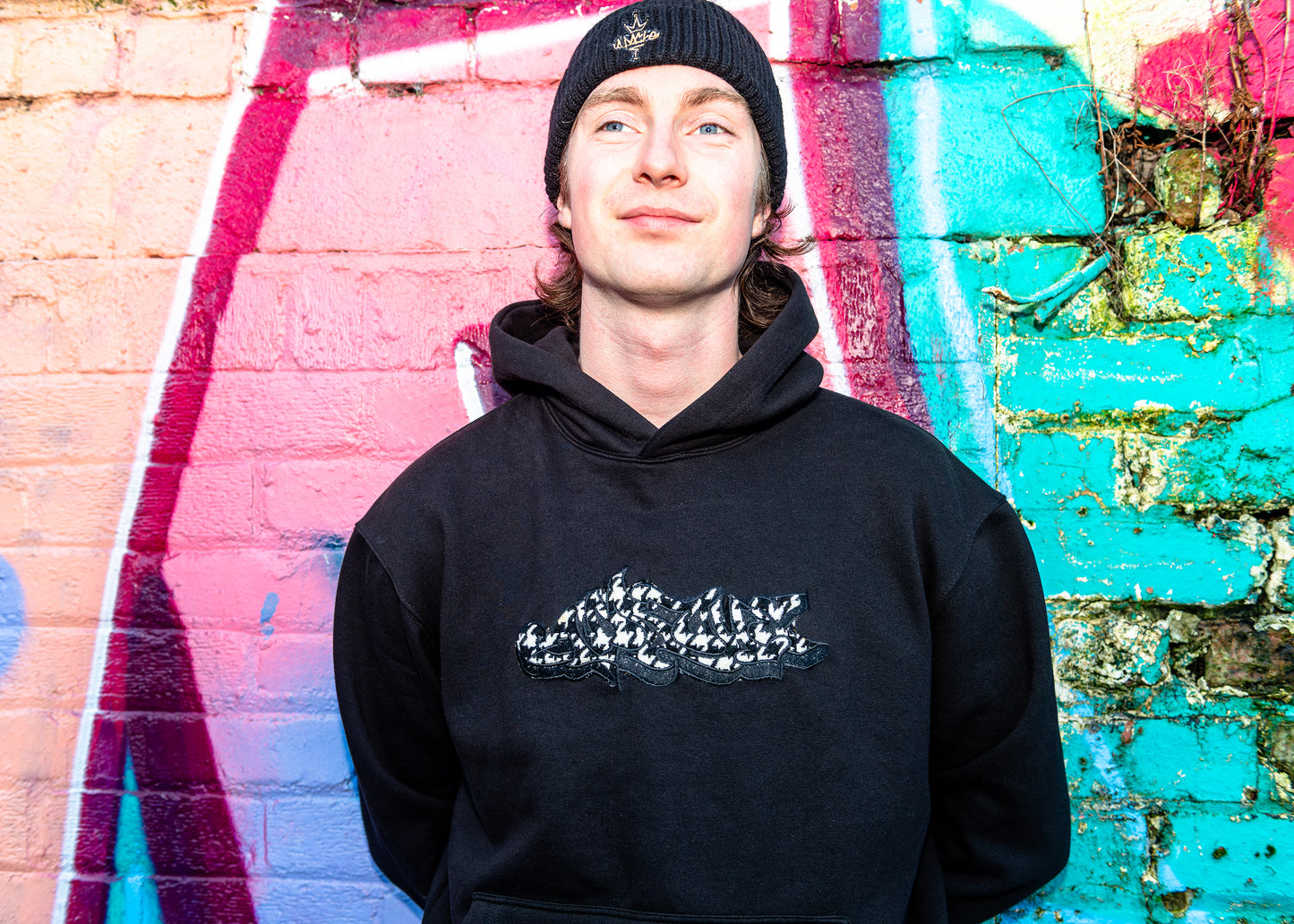 Young man wearing black Heavyweight APACHE Appliqué hoodie with white and black graffiti-style logo patch on chest against colorful wall