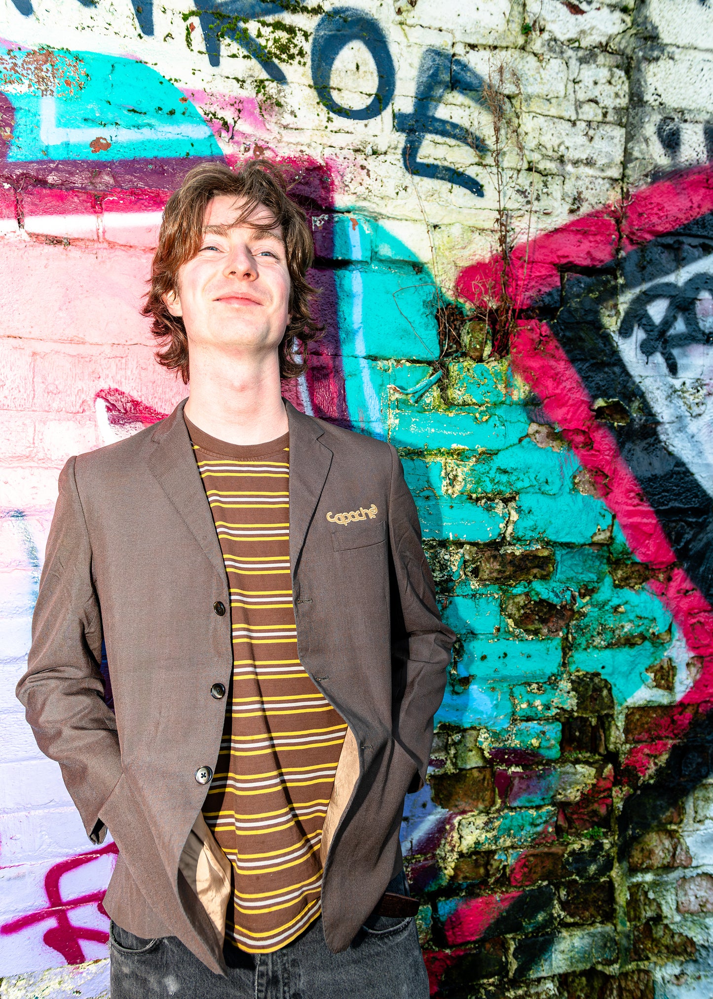 Young man wearing APACHE MEN'S BLAZER in brown with striped shirt, casual streetwear style against colorful graffiti wall