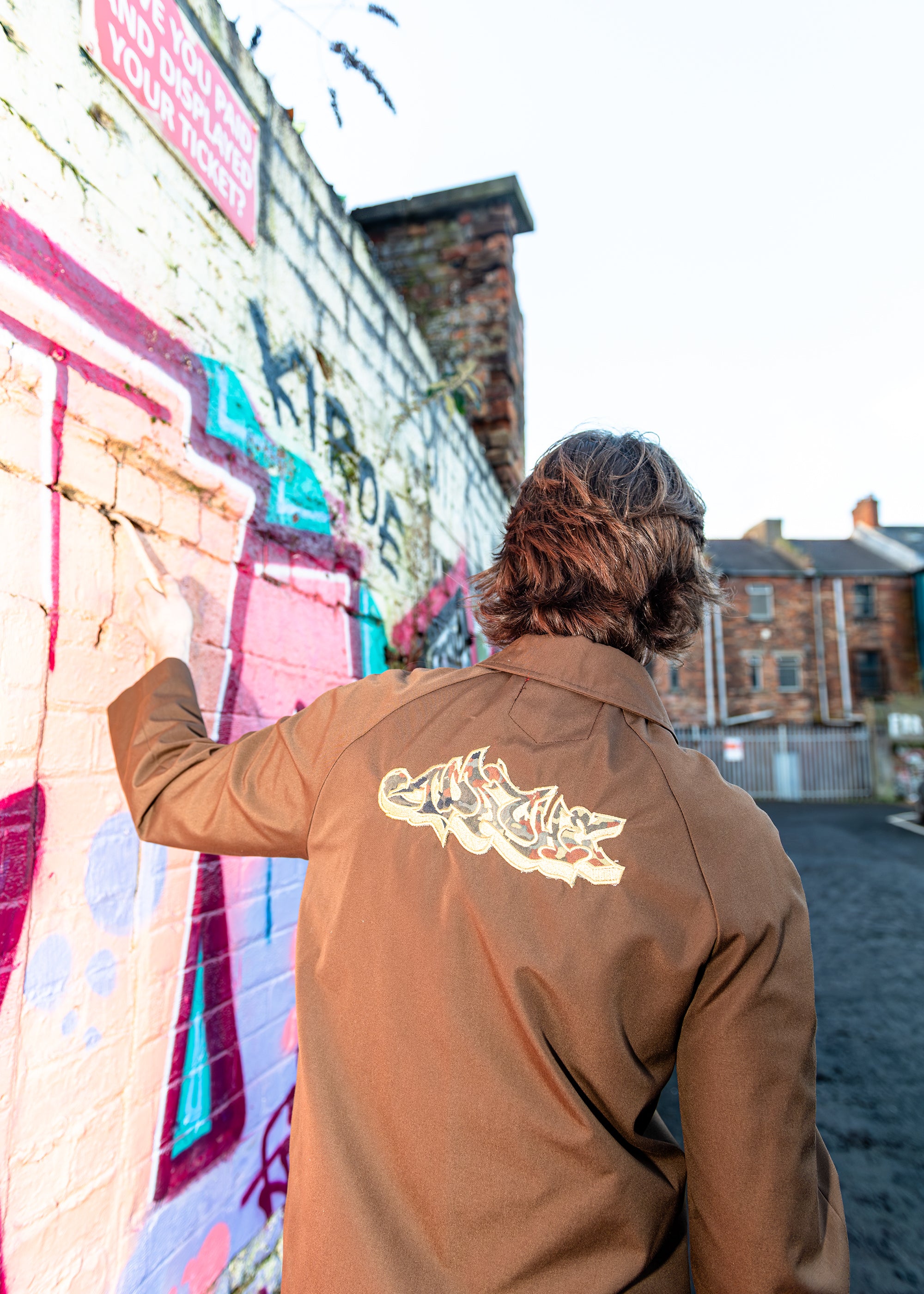 Person wearing Apache Brown/Gold Zip Jacket with gold graffiti-style back print, brown streetwear jacket shown from behind against urban wall