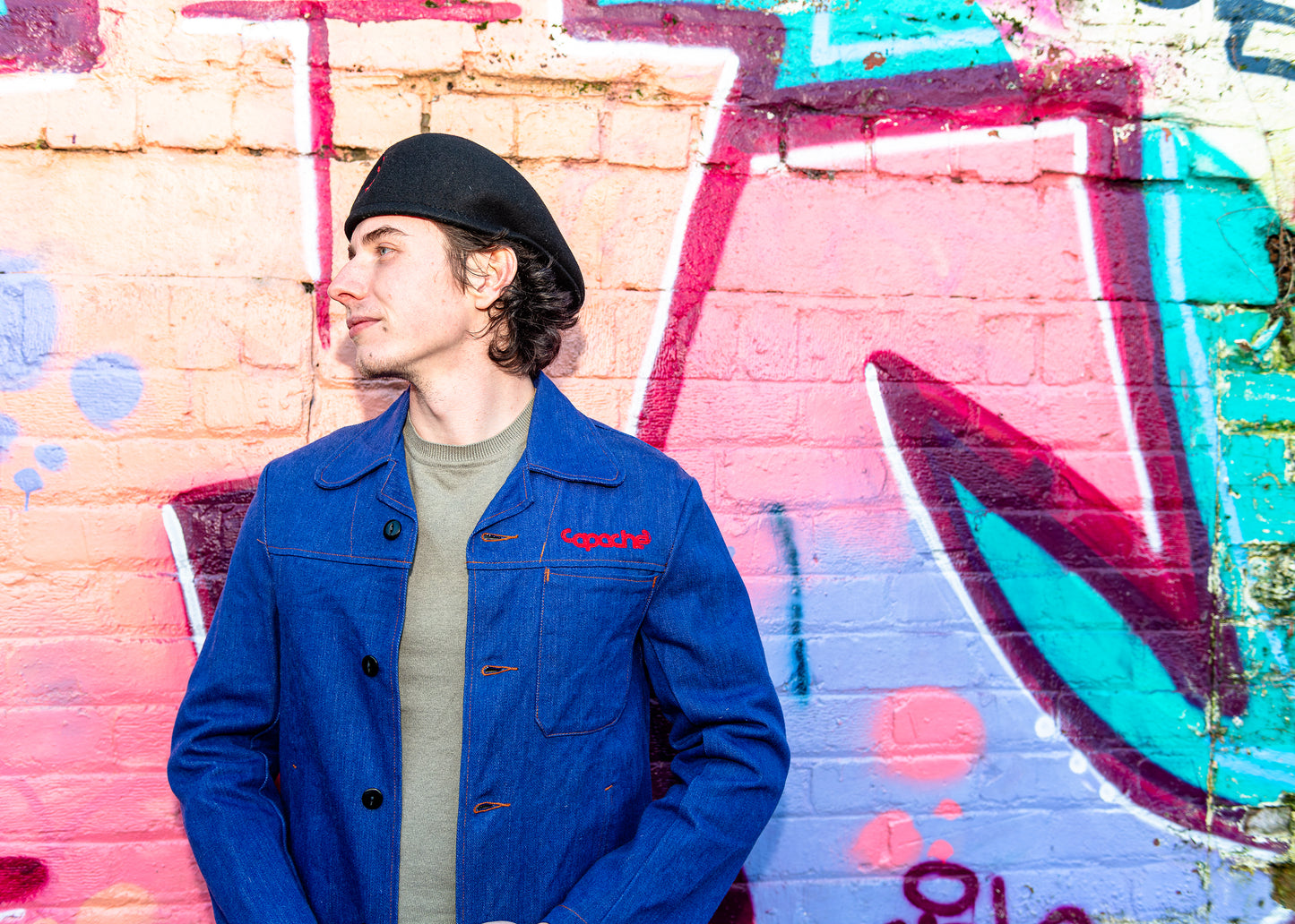 Young man wearing Apache Prison Officer Jacket in blue denim with red Copache embroidery, olive sweater and black beret against colorful graffiti wall