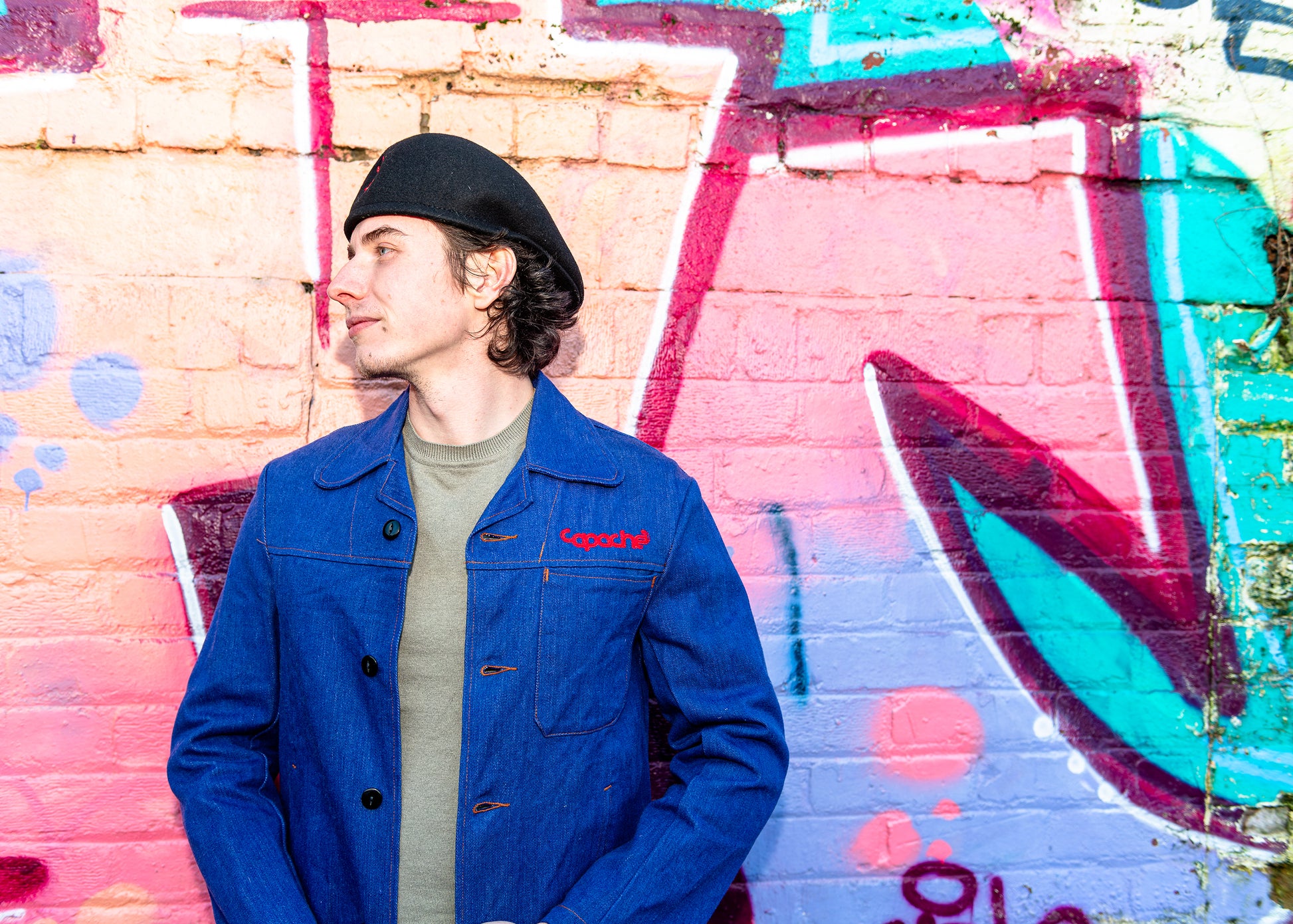 Young man wearing Apache Prison Officer Jacket in blue denim with red Copache embroidery, olive sweater and black beret against colorful graffiti wall