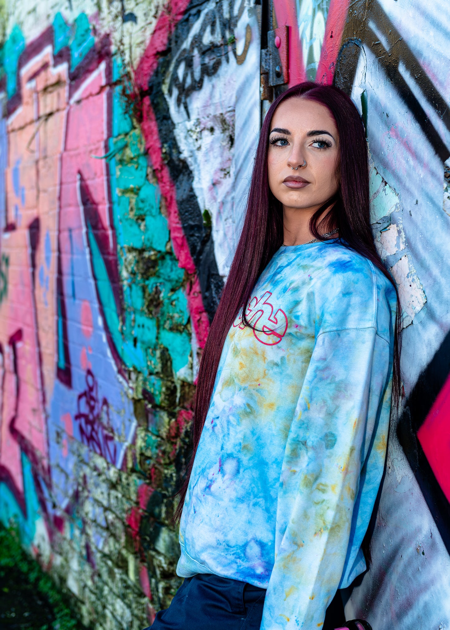 Apache Ice Dye Crewneck in pastel blue tie-dye pattern with pink logo graphic, long sleeve sweatshirt modeled against graffiti wall