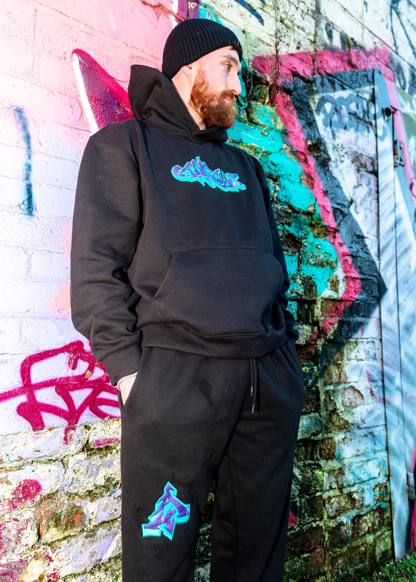 Man wearing black Heavyweight A Joker Appliqué Sweats tracksuit with teal and purple graffiti-style logo on hoodie and joggers