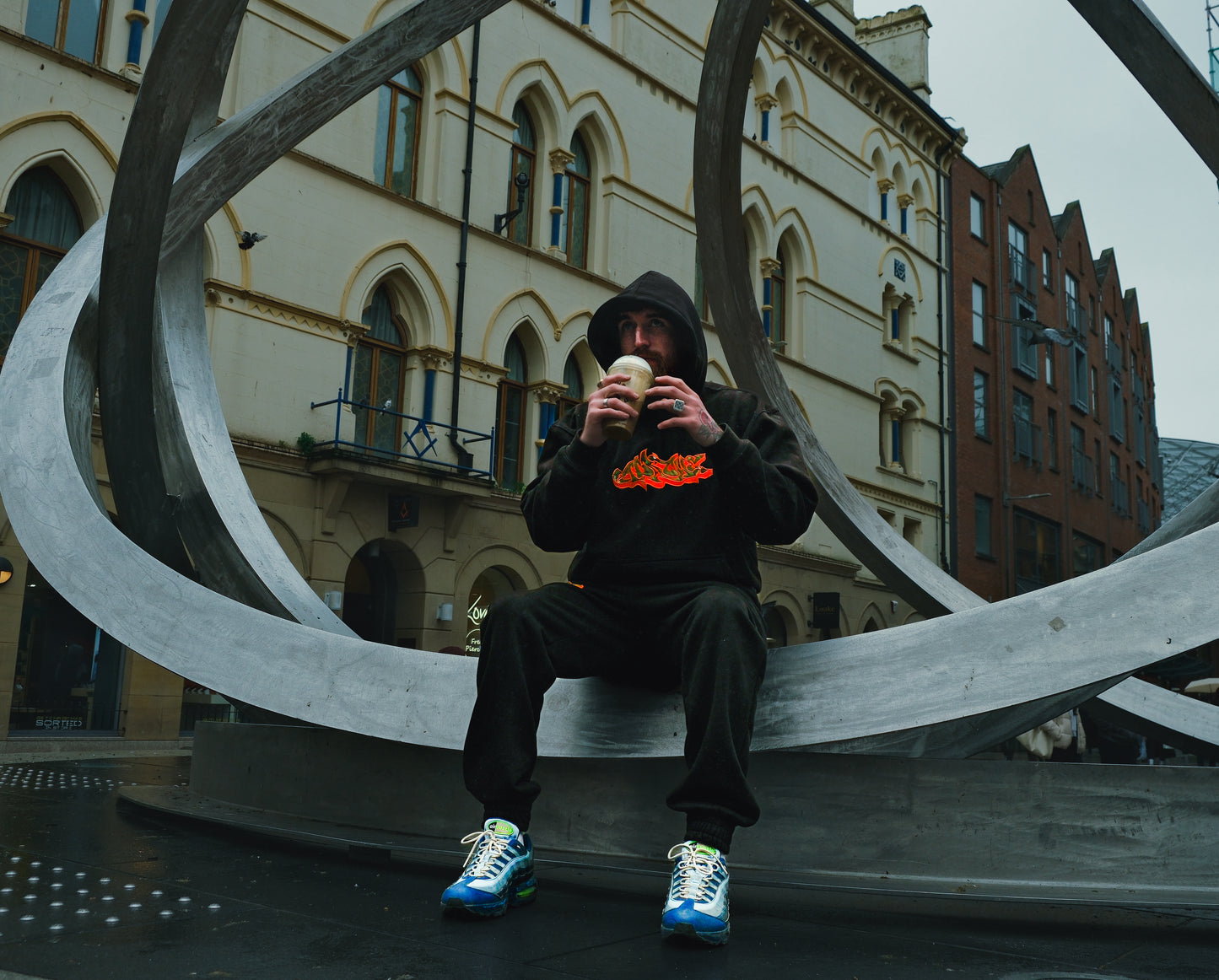 Man wearing black Apache Charizard Appliqué Hoodie with orange flame graphic, sitting on metal sculpture in urban setting