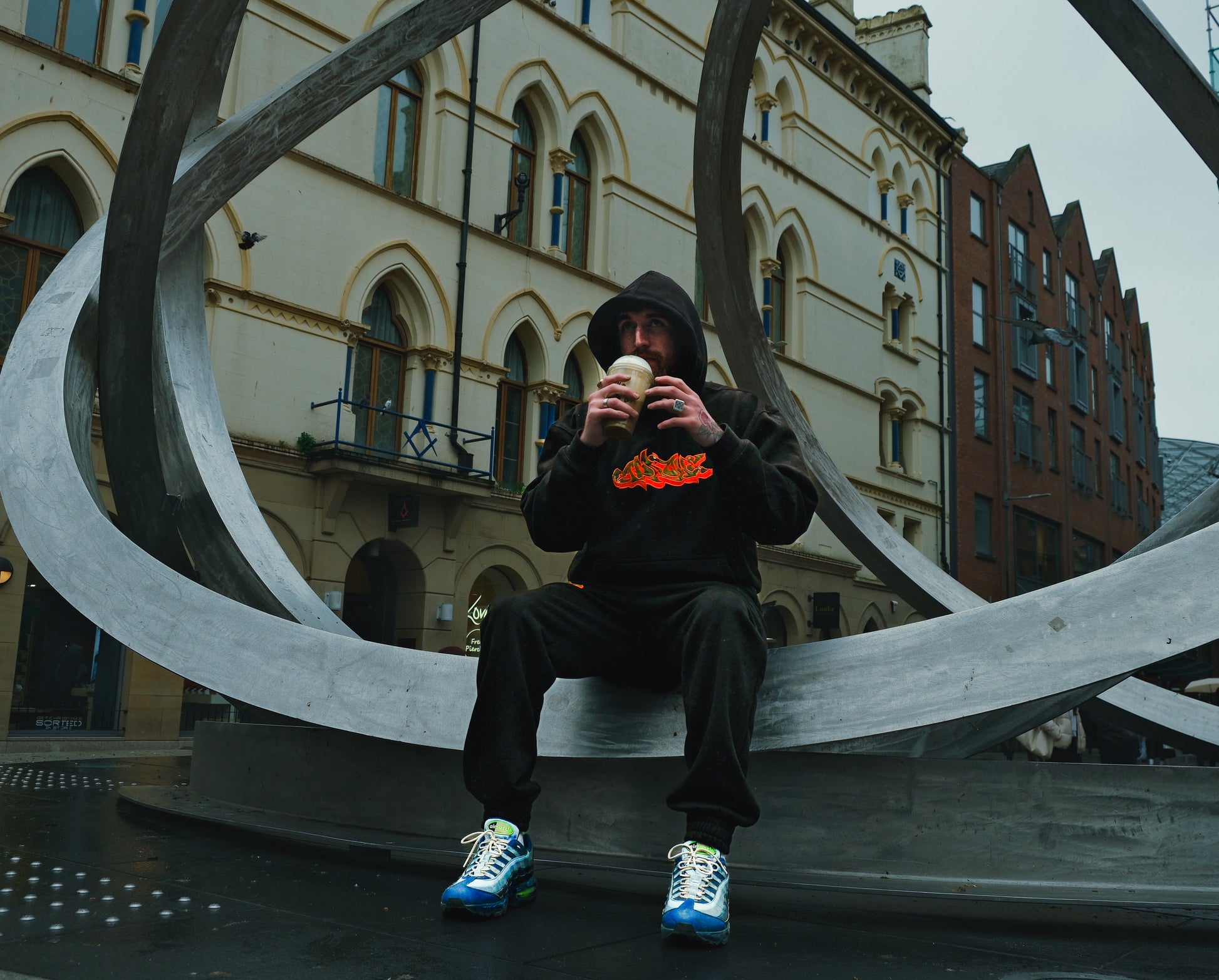 Man wearing black Apache Charizard Appliqué Hoodie with orange flame graphic, sitting on metal sculpture in urban setting
