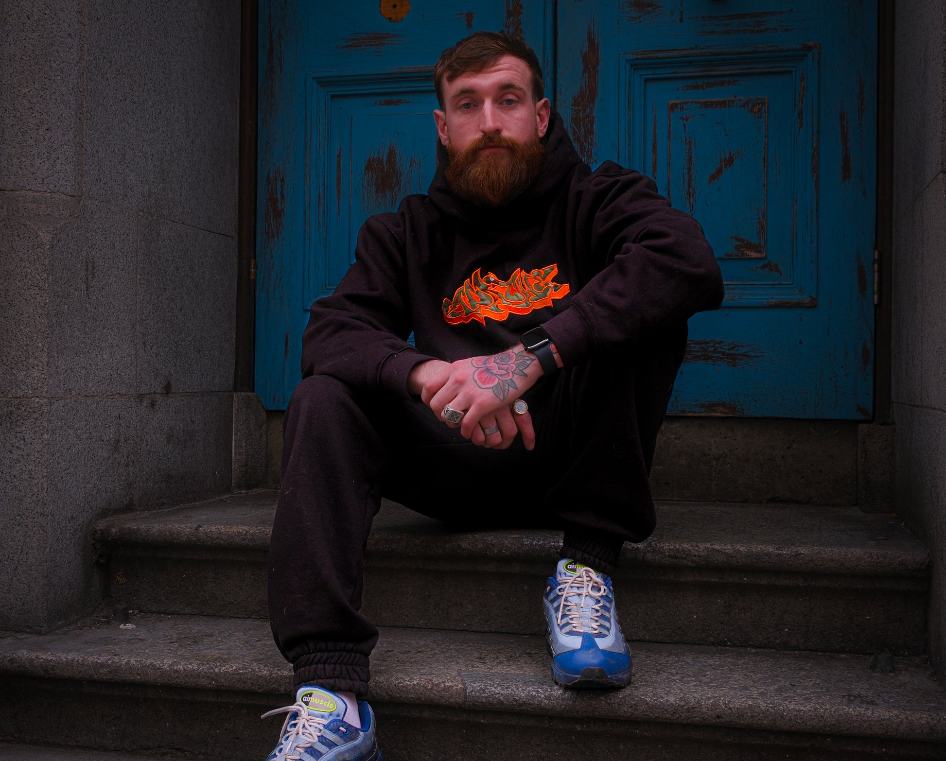 Man wearing black Apache Charizard Appliqué Hoodie with orange graffiti-style graphic, seated on stone steps with blue door backdrop